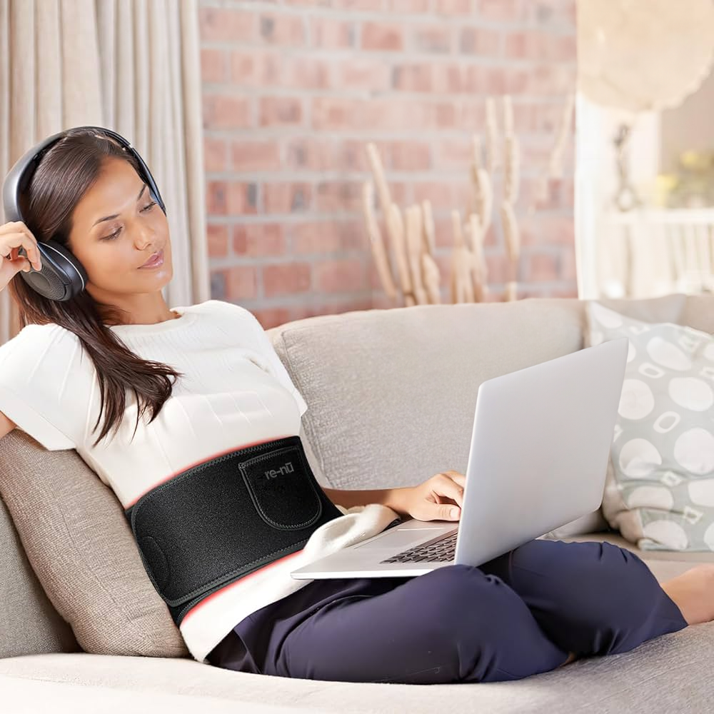 Woman sitting on a sofa, working on her laptop and listening to music with headphones, while comfortably using the re-nū 120 LED Red Light Therapy Belt around her waist for muscle care.