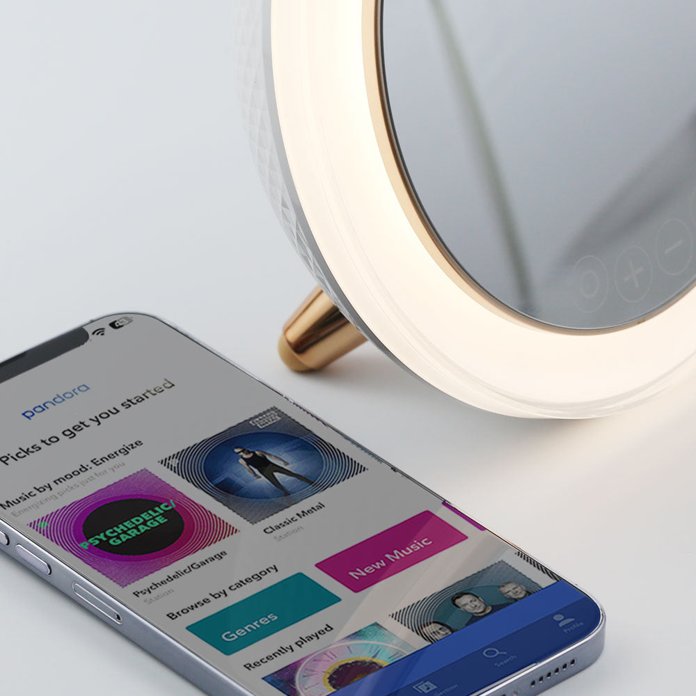 Smartphone placed next to the LED Ring Light Bluetooth Mirror from re-nū Smart Beauty, displaying a music player screen with active song playback to highlight Bluetooth speaker functionality