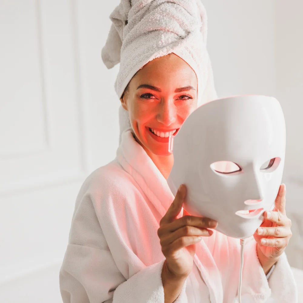 Woman in a bathrobe smiling while holding an LED beauty mask, ready for an advanced skincare light therapy session.