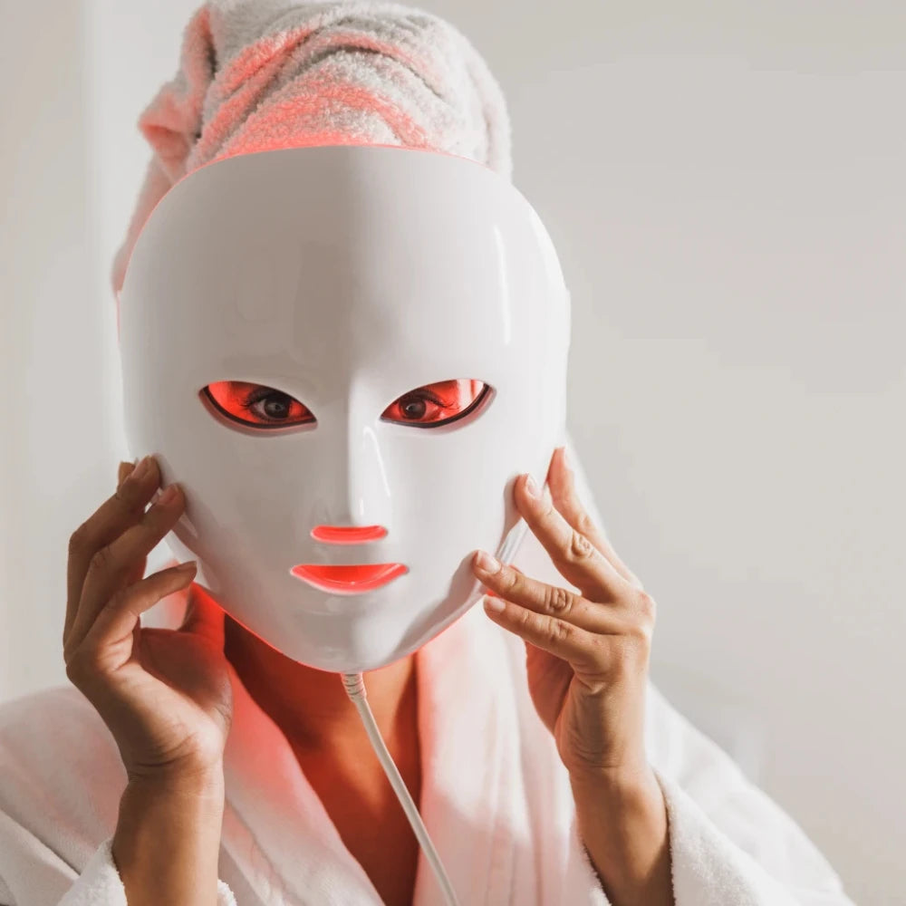 Front-facing view of a woman wearing the re-nū 8 Color LED Facial Mask, gently supporting it with her hands during a light therapy session.