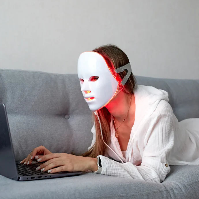 Woman laying on a sofa and working on her laptop while wearing the re-nū 8 Color LED Facial Mask, showing hands-free skincare during a daily routine.