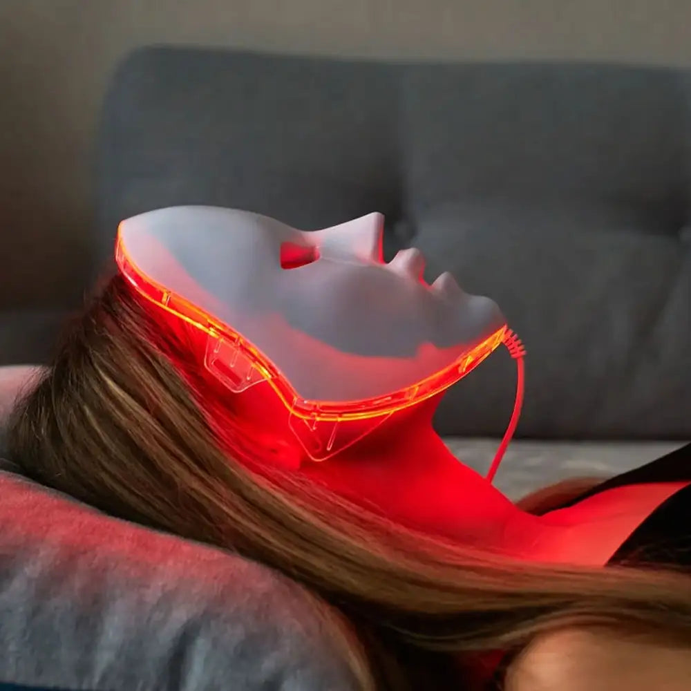 Woman laying on couch relaxing and using a red light therapy mask from the comfort of her own home.