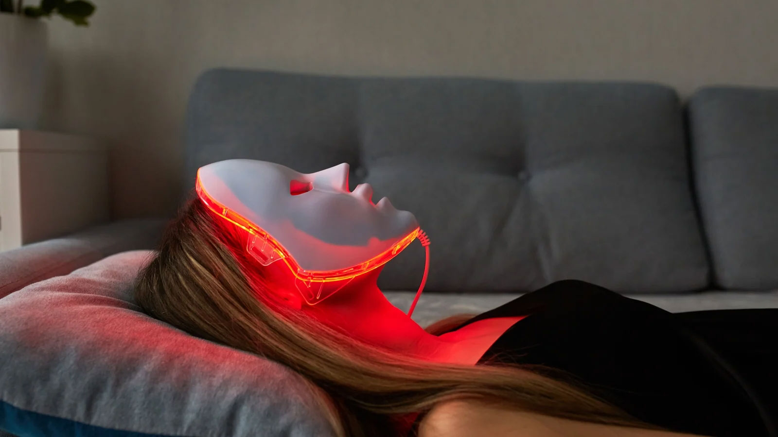 Woman laying on couch relaxing and using a red light therapy mask from the comfort of her own home.
