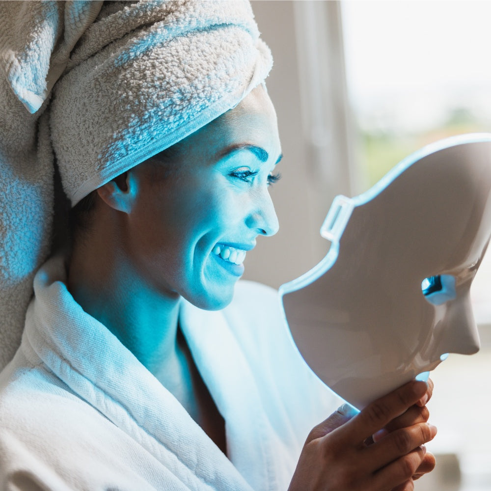 A woman in a bathrobe holds an LED light therapy mask, showcasing a skincare treatment for rejuvenation.