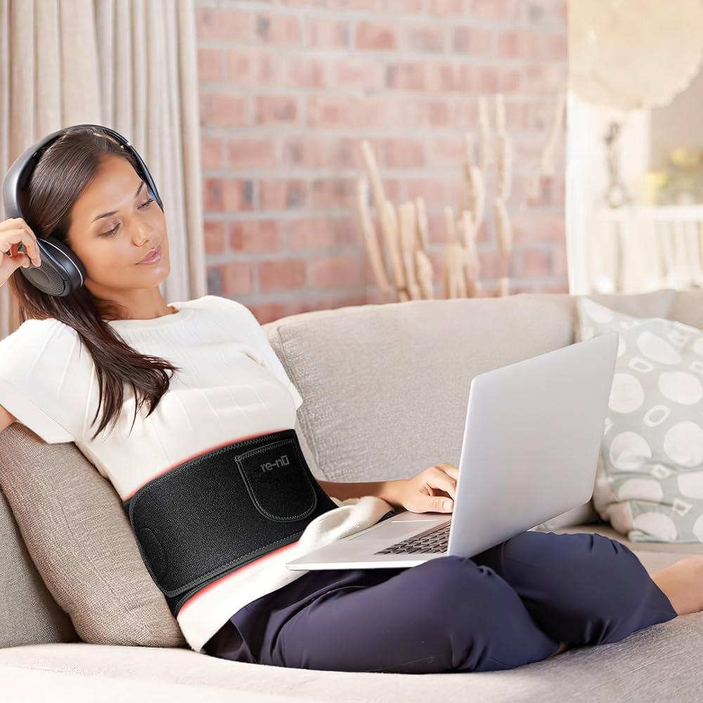 A woman relaxing on a couch with headphones, engaged with her laptop, while a Mask LED Light Therapy device glows.