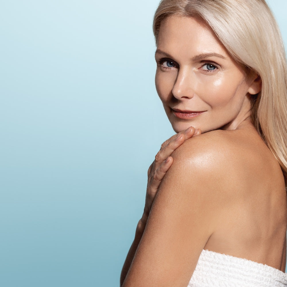 A woman with long blonde hair poses in a towel, showcasing her skin illuminated by a Mask LED Light Therapy device.