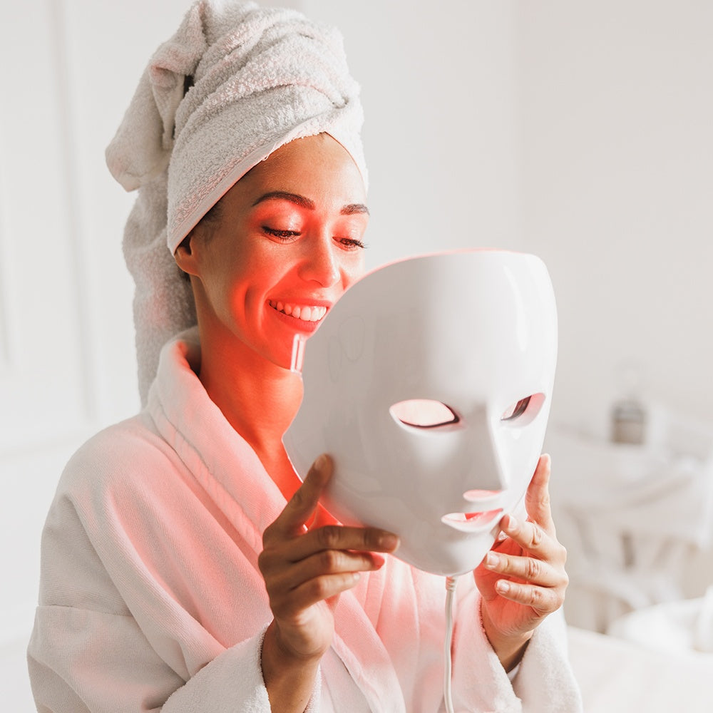 A woman in a bathrobe holds an LED light therapy mask, showcasing a skincare treatment for rejuvenation.