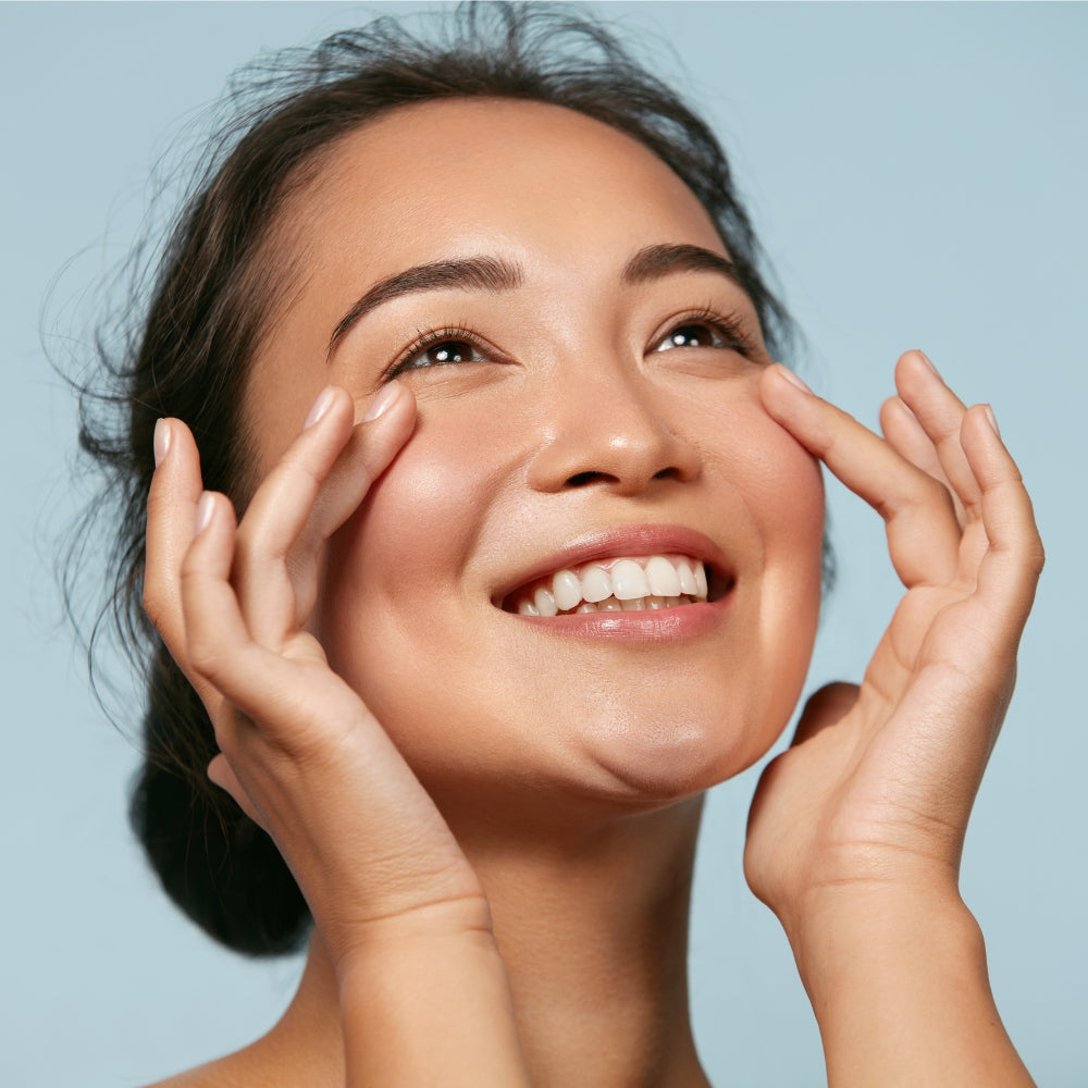 A woman smiling while gently touching her eyes, showcasing the benefits of Mask LED Light Therapy.