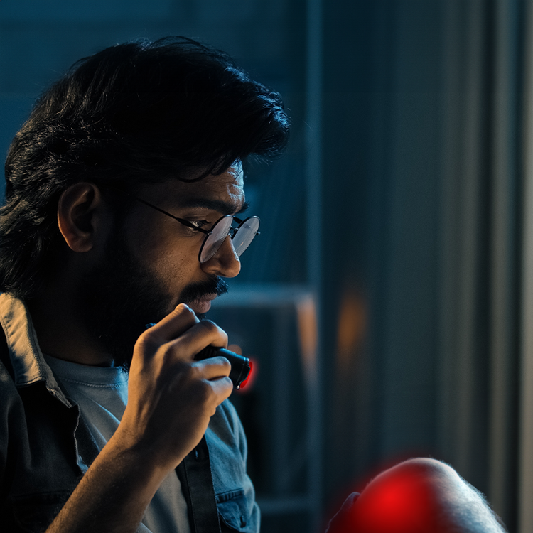 A man with glasses and a beard sits in front of a computer, using a Mask LED Light Therapy device for skin treatment.