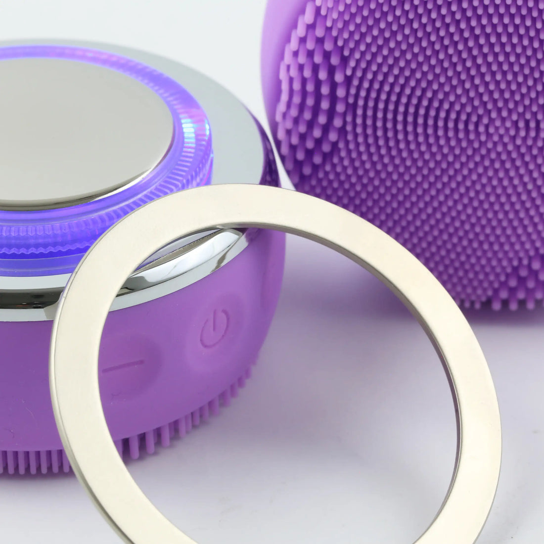 PurePulse Pro Plus Sonic Cleansing Brush illuminated with purple LED resting face down with stainless steel magnetic ring attachment displayed beside the device