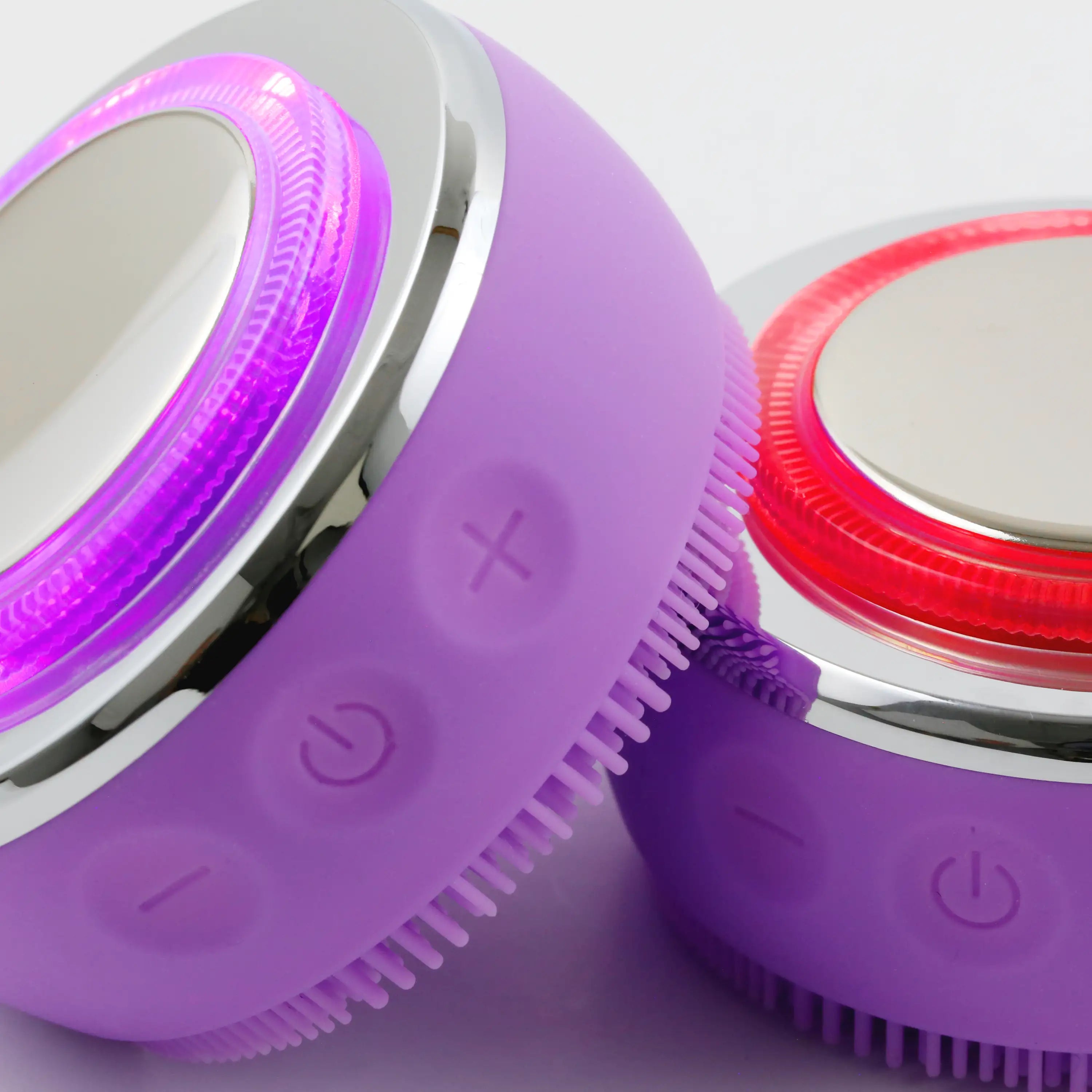 Close-up of the PurePulse Pro Plus chrome EMS head with red LED illumination beside a second brush resting on its side with purple LED glow and reflective metallic detailing