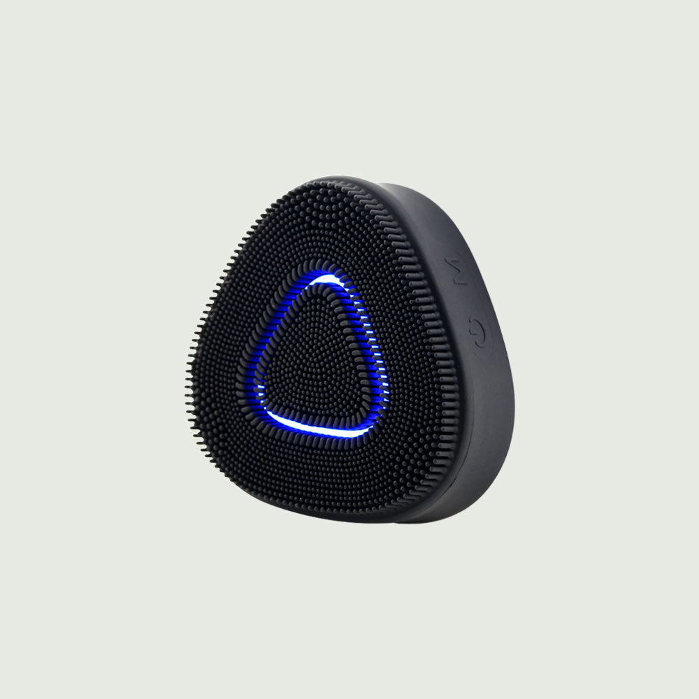 Close-up of a black triangular sonic cleansing brush featuring soft silicone bristles and a glowing blue LED light ring, designed for deep facial cleansing and exfoliation