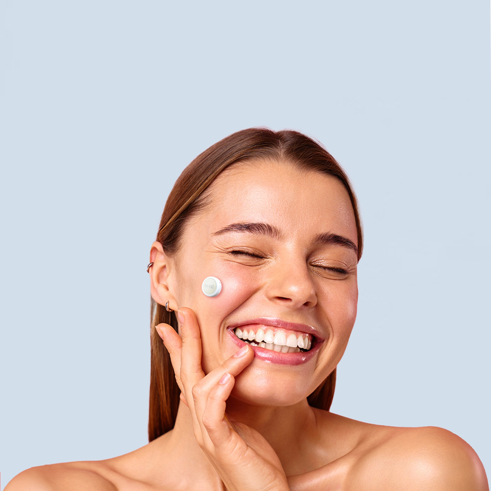 A woman smiles as she applies a cream to her face, showcasing the benefits of LED light therapy for skin rejuvenation.