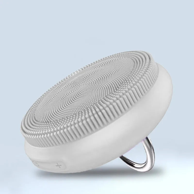 A compact white silicone facial cleansing brush with textured bristles and a silver ring handle, displayed on a blue background for deep pore cleansing and gentle exfoliation.