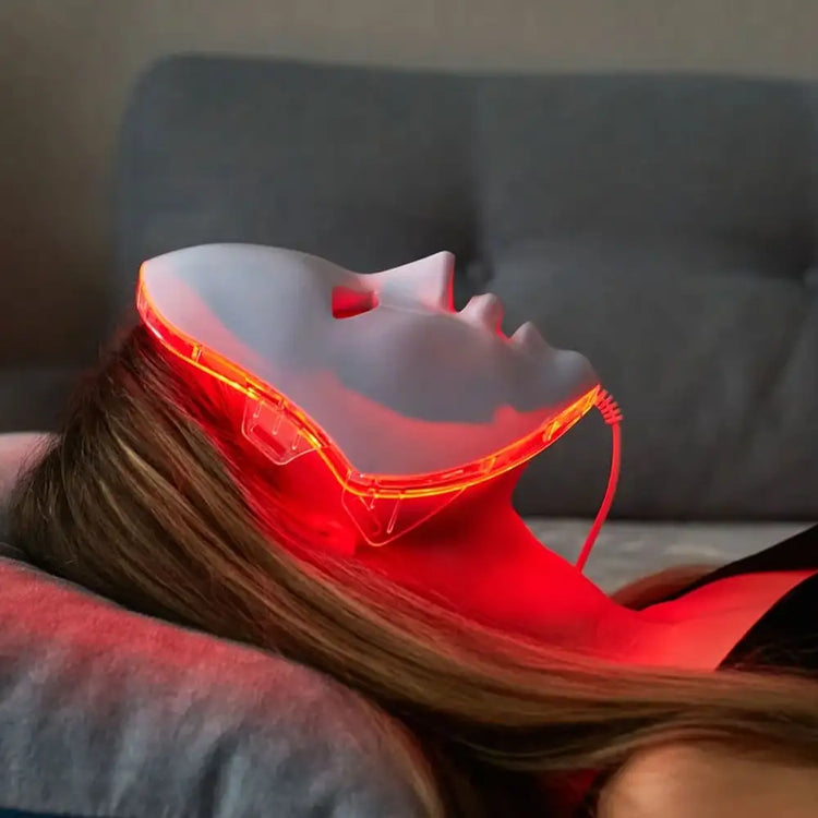 Woman laying on couch relaxing and using a red light therapy mask from the comfort of her own home.