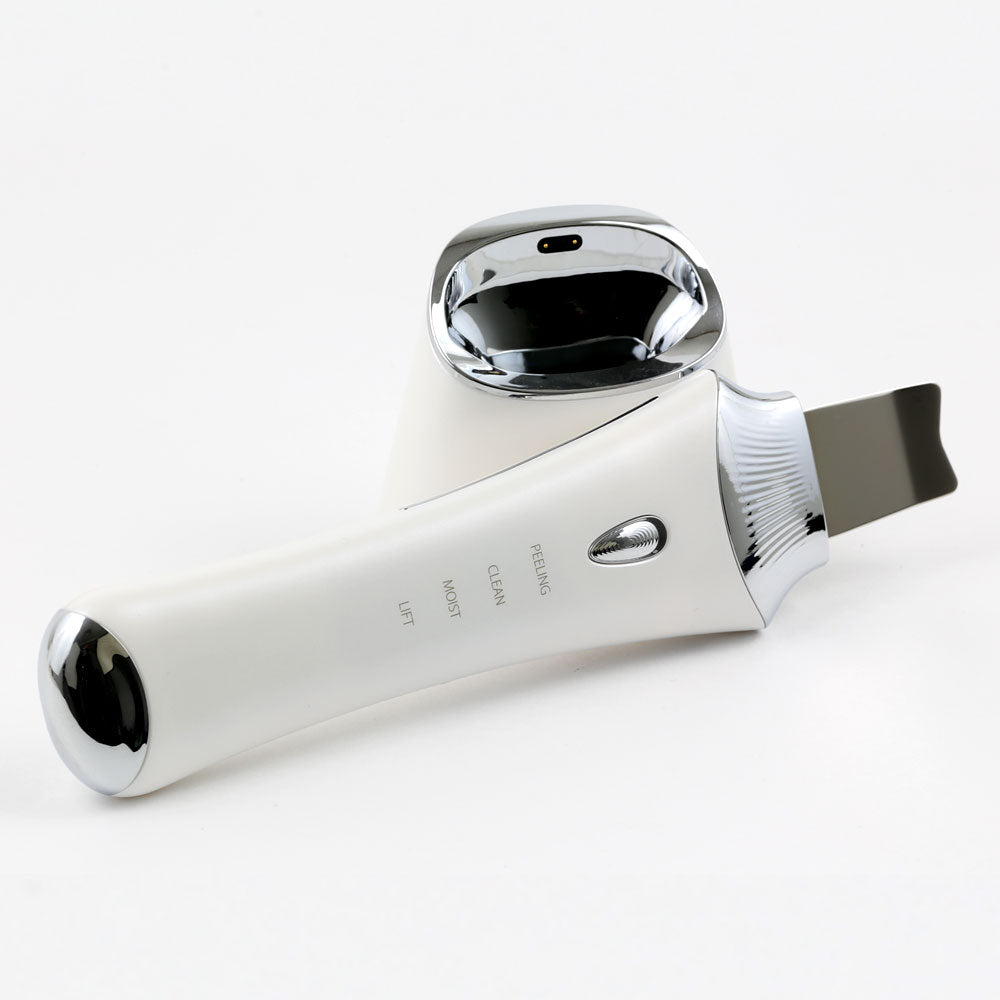 Tabletop side view of the Ultrasonic Skin Scrubber with UV Base, highlighting its wireless UV charging base, easy to use power/mode button; and sleek stainless EMS spatula.