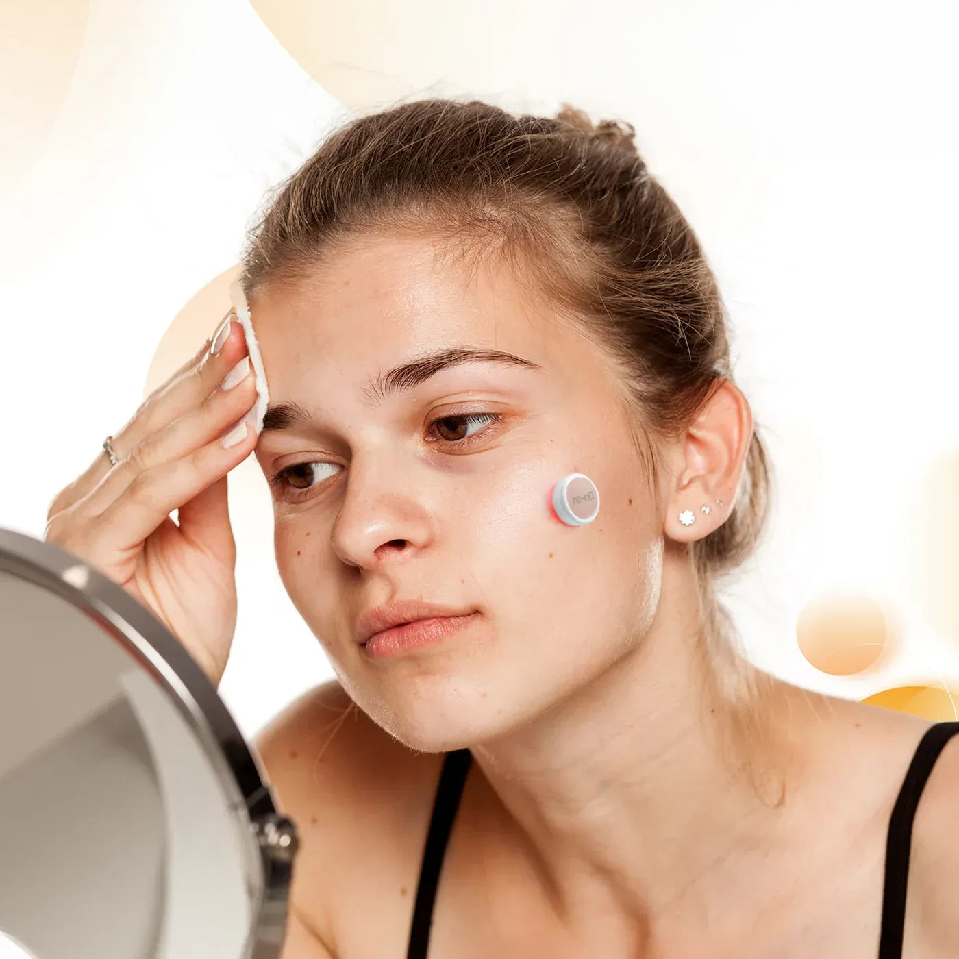 Woman with beautiful skin in front of a mirror cleansing her face with a pad while wearing the GlowUp Acne LED Patch on her left cheek for acne treatment.