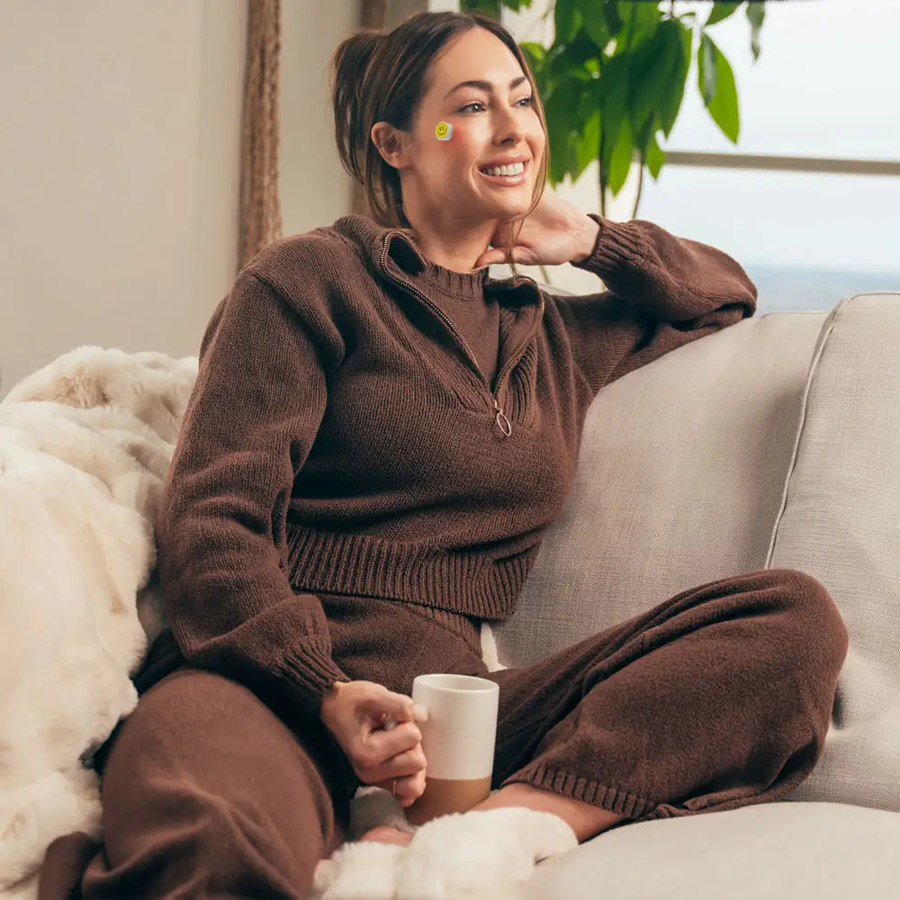 Beautiful smiling woman sitting on a couch with a cup of coffee, wearing the Smart LED Recovery Patch on her right cheek for red and near-infrared light acne treatment.