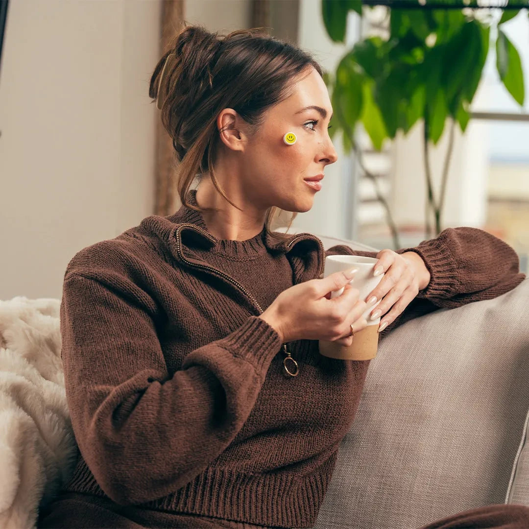 Beautiful woman sitting on a couch with a cup of coffee, wearing the GlowUp Acne LED Patch on her right cheek for blue light acne treatment.